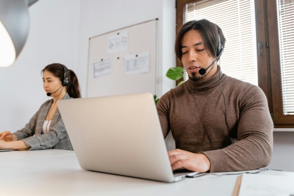 Two call center employees working together, wearing headsets, in an office setting.